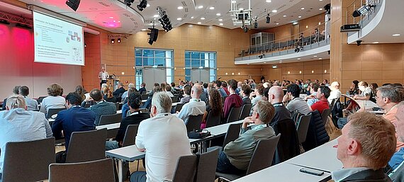 Bild einer Konferenz mit einem Zuschauerraum von hinten und Blick aufs Podium mit Sprecher.